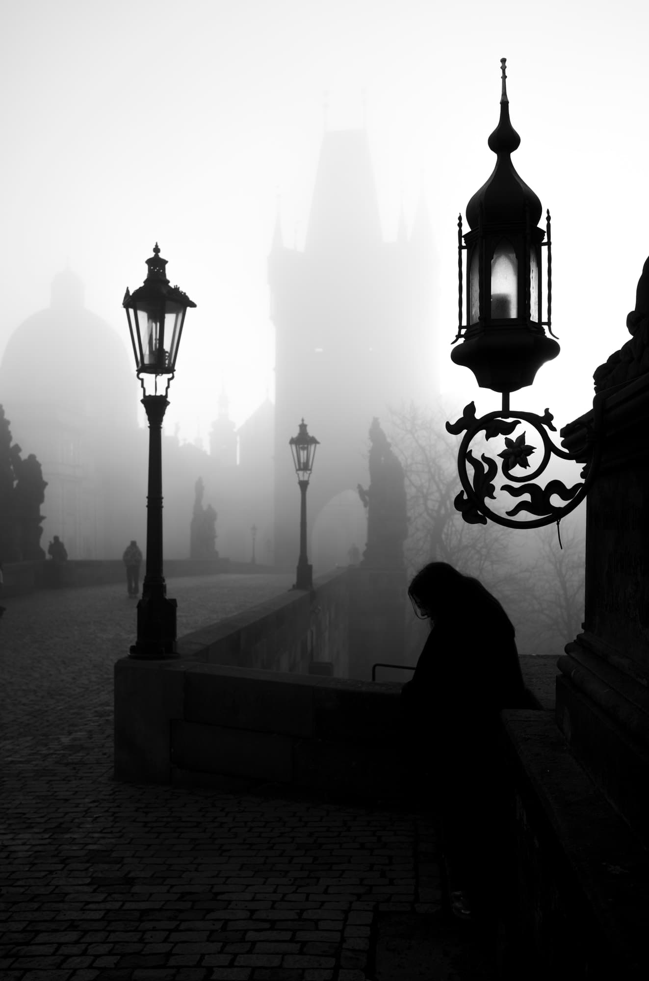 Ornate gas lanterns on Charles Bridge in dense fog, a solitary figure leaning on the parapet